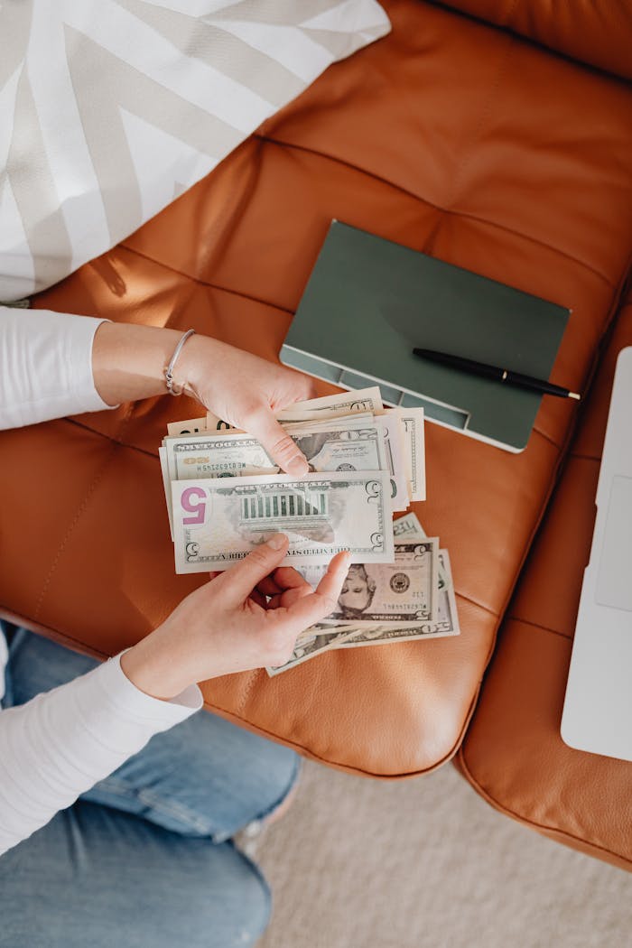 Person counting US dollar bills on a leather sofa with a notebook nearby.